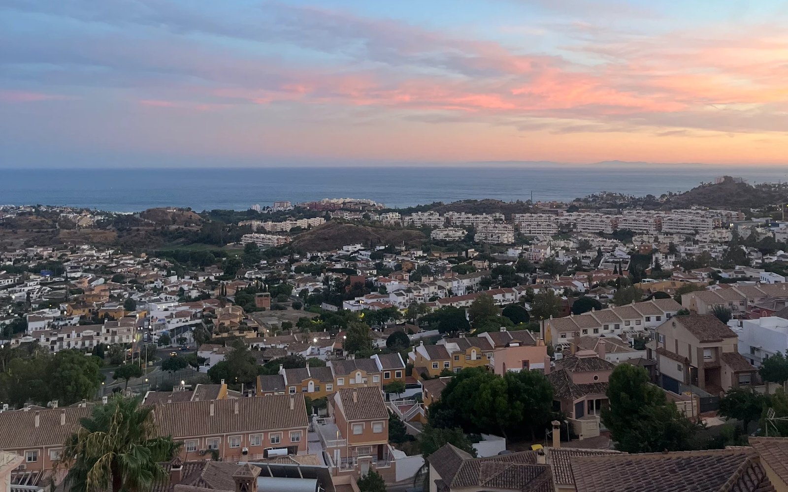 Vista al atardecer desde una urbanización en las alturas de la Costa del Sol, con tejados, el mar y la luna en el cielo rosado